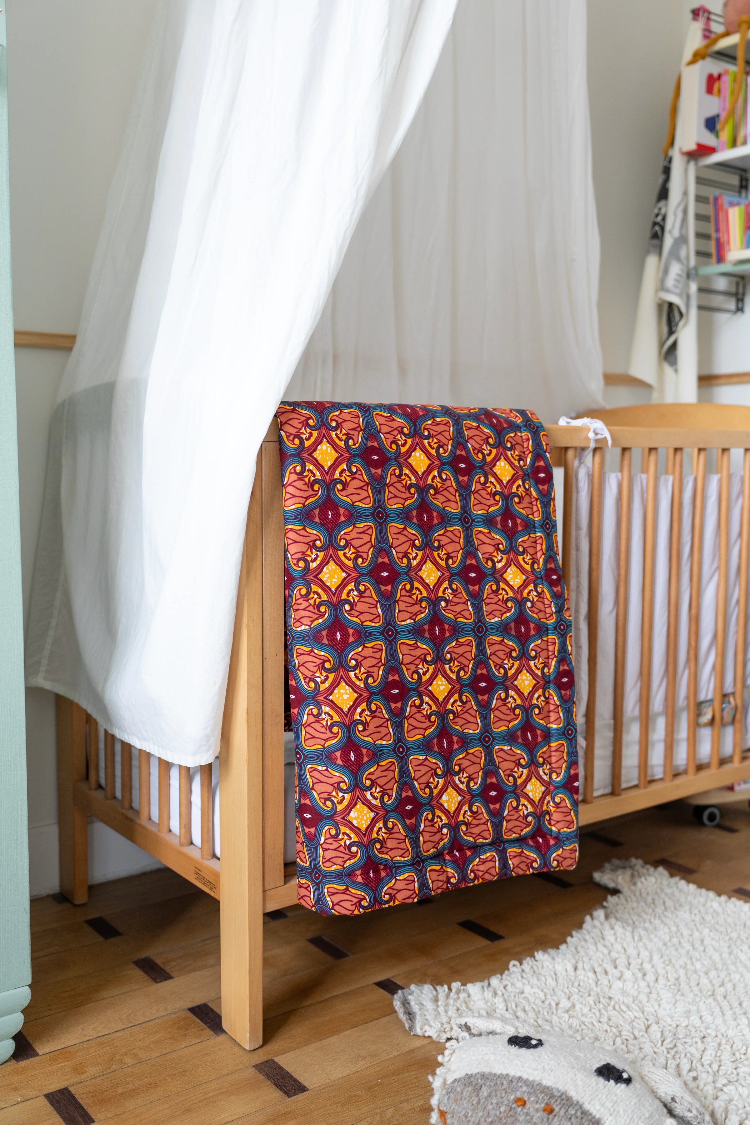 Dans une chambre bébé, édredon bébé Malaika en wax et double gaze de coton bois de rose et bordeaux, posé sur le bord d'un lit à barreaux. Au sol, le tapis mouton SHEEPY en pure laine de Studio Matongé