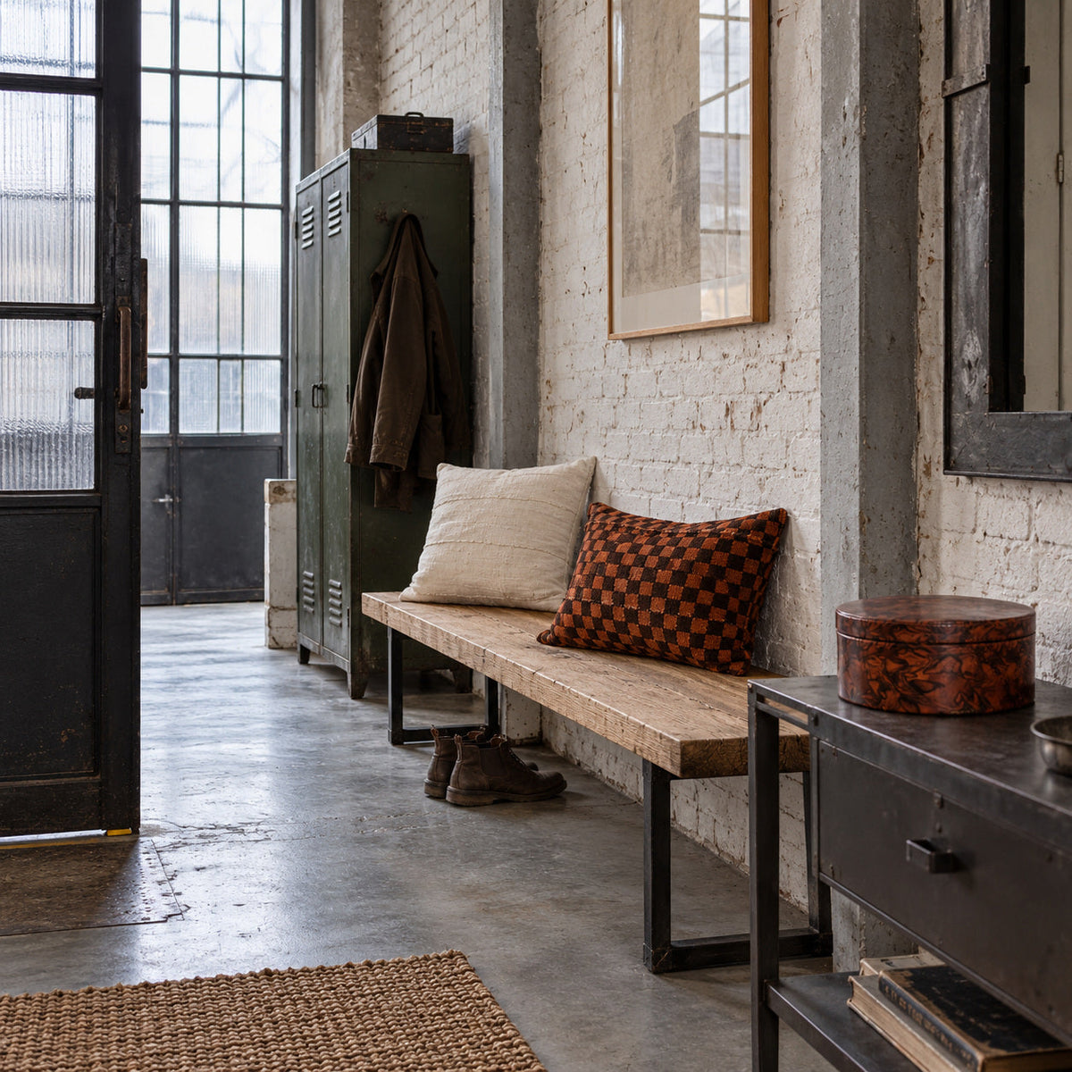 Entrée industrielle avec coussins TANDA terracotta et MSINGI écru sur un banc, et boîte DEÑC-MITI sur une console.