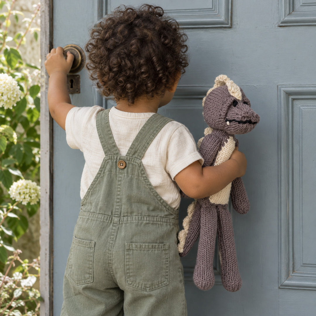 Enfant en salopette sauge tenant le doudou crocodile MAXIME taupe devant une porte bleu grisé.