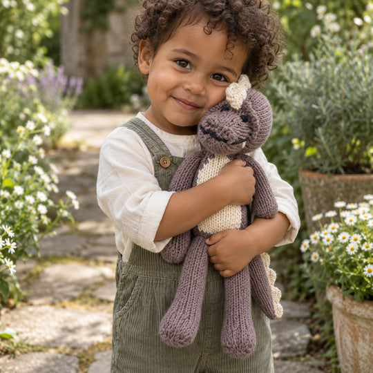 Enfant aux cheveux bruns bouclés câlinant le doudou crocodile MAXIME taupe dans un jardin.
