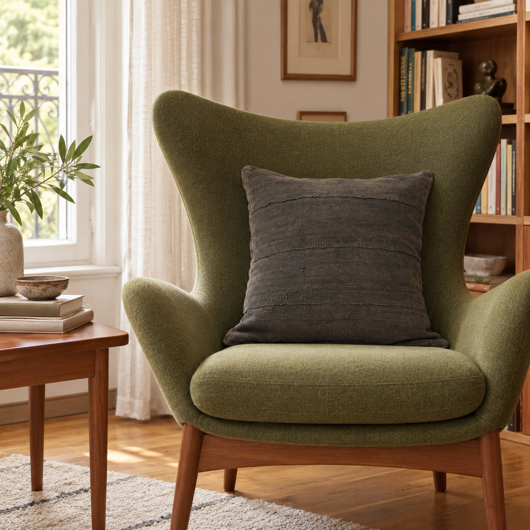 Vierkant MSINGI Encre kussen van zwarte bogolan op een olijfgroene fauteuil, bij een raam en een houten boekenkast.
