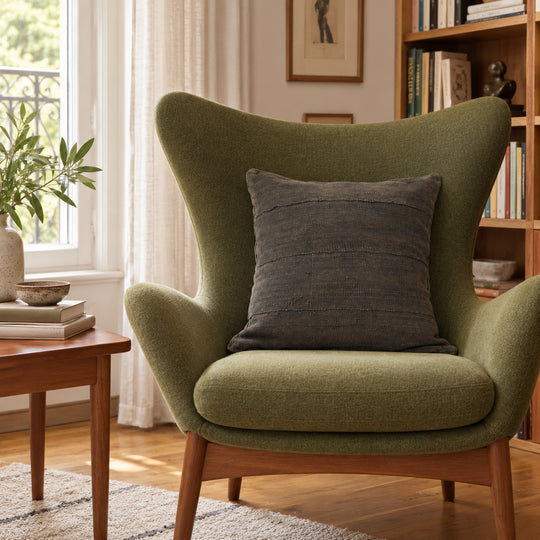 Vierkant MSINGI Encre kussen van zwarte bogolan op een olijfgroene fauteuil, bij een raam en een houten boekenkast.