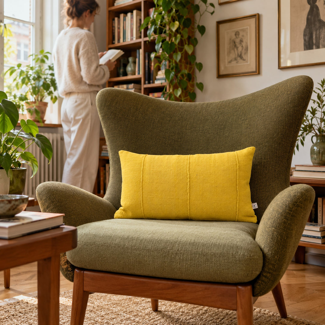 Mimosageel MSINGI kussen op een olijfgroene fauteuil, naast een boekenkast en een persoon die door een boek bladert.
