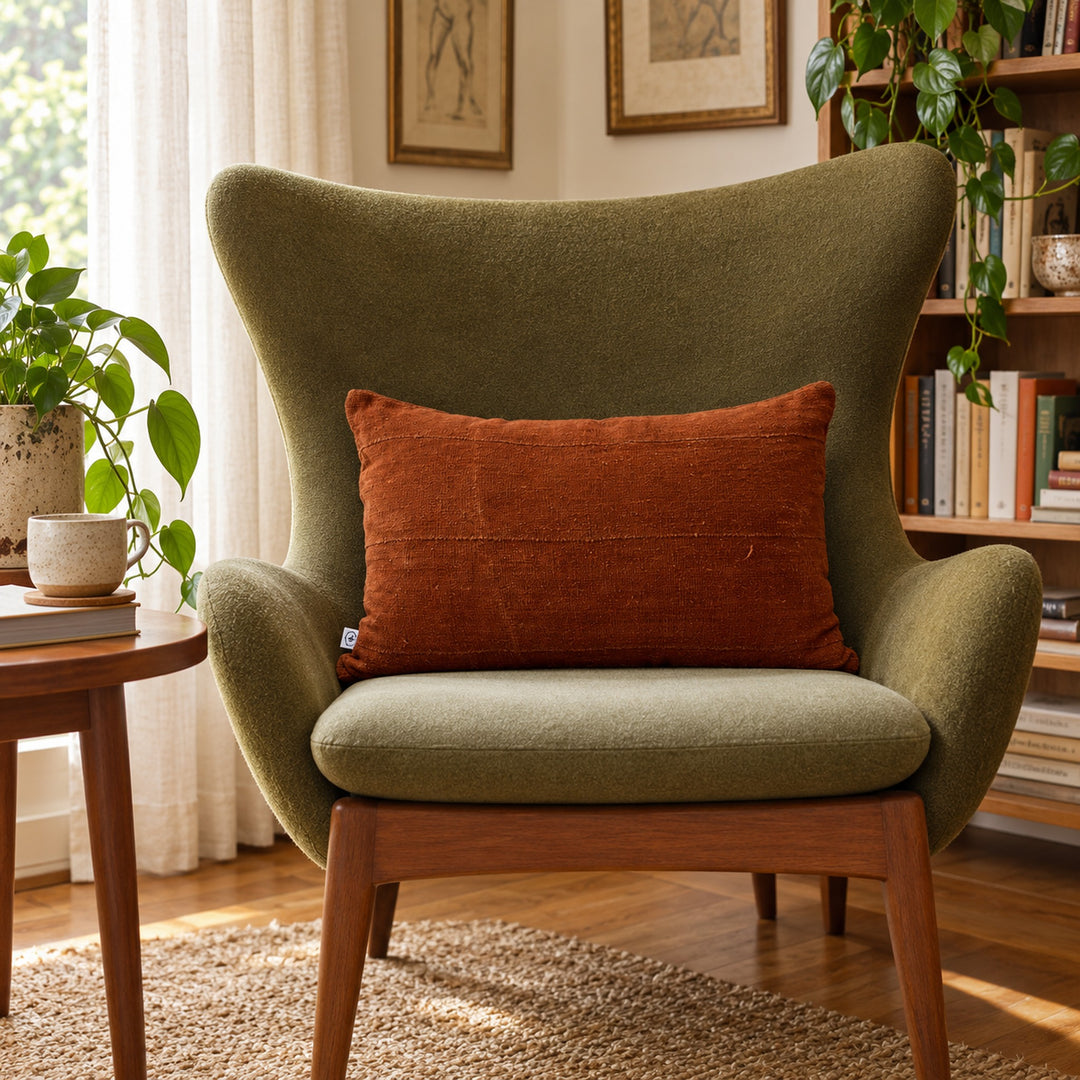 Rechthoekig terracottakleurig MSINGI kussen op een olijfgroene fauteuil, naast een houten tafel met een boek en een kopje.