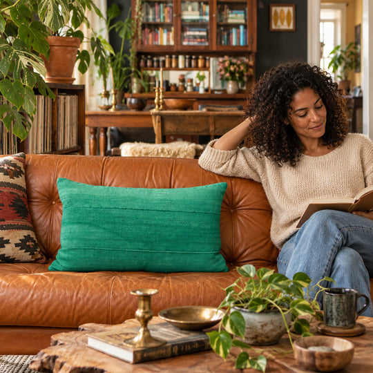 Rechthoekig groen MSINGI kussen op een karamelkleurige leren bank, in een woonkamer met boekenkast, planten en een zittende persoon.