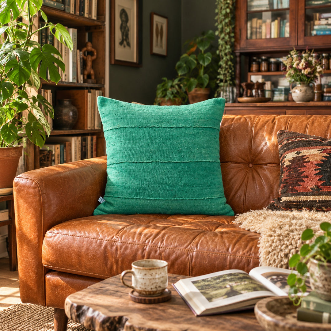 Vierkant groen MSINGI kussen op een karamelkleurige leren bank, in een woonkamer met een houten boekenkast, planten en een opengeslagen boek.