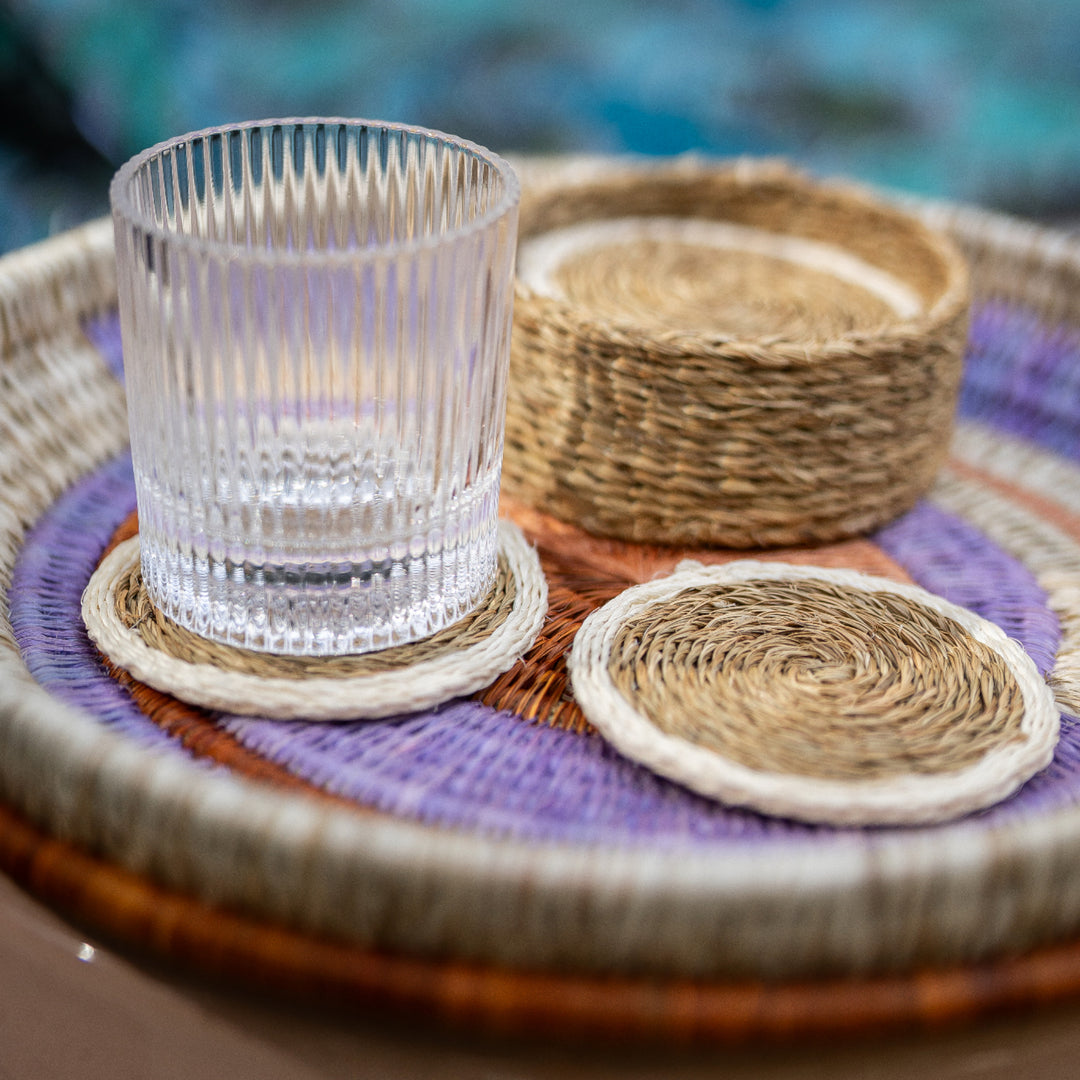 Sous-verres LUTINDZI en fibres naturelles avec verre transparent et panier assorti, mis en scène sur un plateau rond tressé aux tons crème, parme et terracotta.