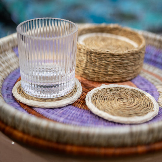 Sous-verres LUTINDZI en fibres naturelles avec verre transparent et panier assorti, mis en scène sur un plateau rond tressé aux tons crème, parme et terracotta.