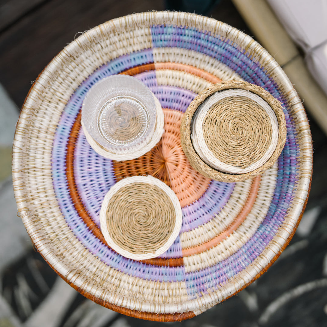 Vue de dessus de sous-verres LUTINDZI en fibres naturelles avec verre transparent et panier assorti, présentés sur un plateau rond tressé aux tons doux.
