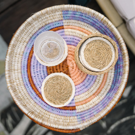 Vue de dessus de sous-verres LUTINDZI en fibres naturelles avec verre transparent et panier assorti, présentés sur un plateau rond tressé aux tons doux.