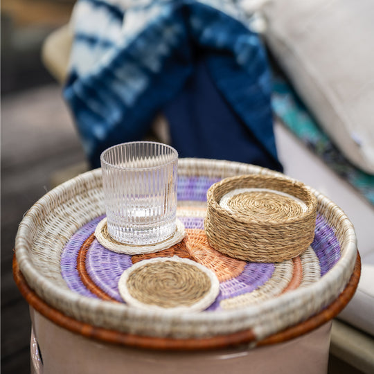 Sous-verres LUTINDZI en fibres naturelles avec verre transparent et panier assorti, disposés sur un plateau rond tressé multicolore dans un salon.