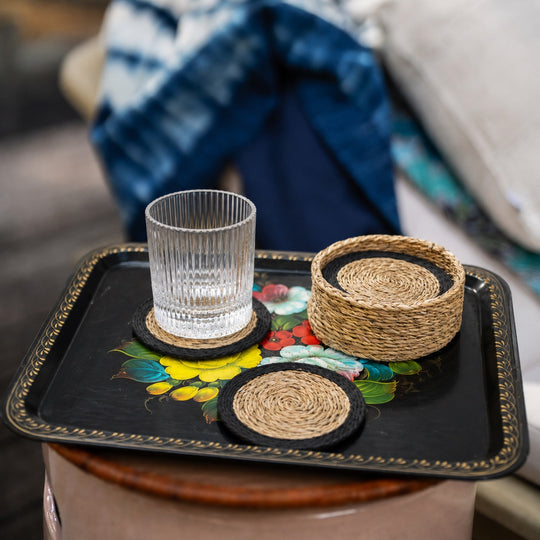 Sous-verres tressés en fibres naturelles avec bord noir et panier assorti, posés sur un plateau métallique vintage à décor floral, avec verre transparent.