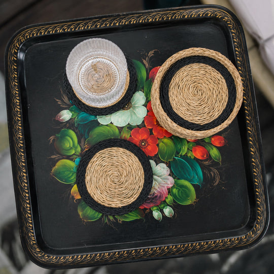 Vue de dessus de sous-verres tressés en fibres naturelles avec bord noir et panier assorti, disposés sur un plateau métallique noir à motif floral vintage avec verre transparent.