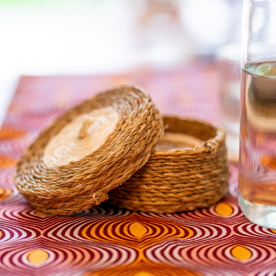 Gros plan sur un panier à sous-verres LUTINDZI en fibres naturelles, posé sur une nappe en wax graphique rose, orange et bordeaux, avec verre transparent.