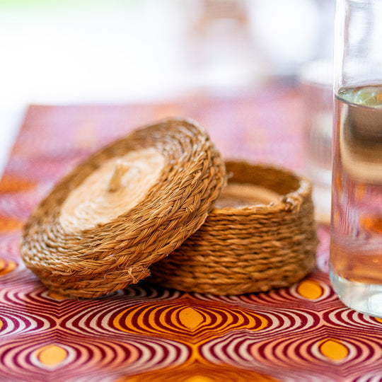 Gros plan sur un panier à sous-verres LUTINDZI en fibres naturelles, posé sur une nappe en wax graphique rose, orange et bordeaux, avec verre transparent.