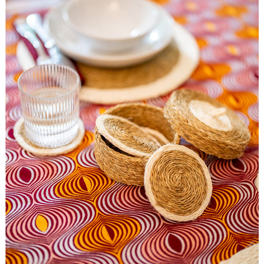 Sous-verres LUTINDZI en fibres naturelles avec panier assorti et verre strié, disposés sur une nappe en wax rose, orange et bordeaux.
