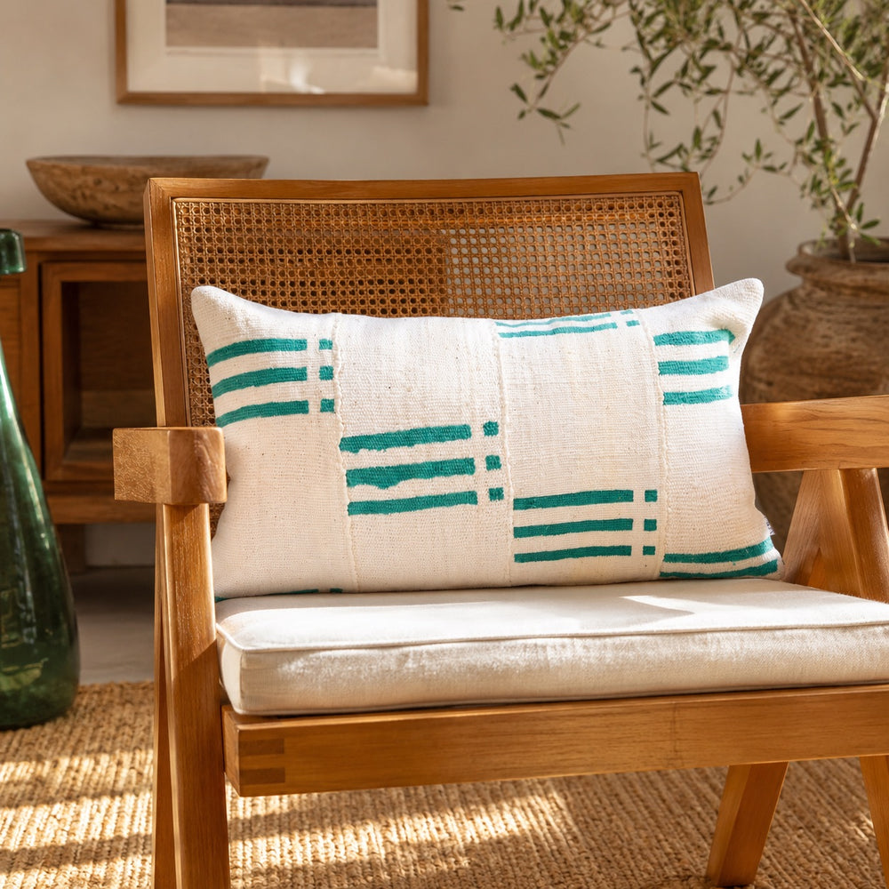 Housse de coussin NZETE par Studio Matongé en bogolan blanc et vert, posée sur un fauteuil en bois et cannage dans un salon lumineux à l'esprit seventies. 
