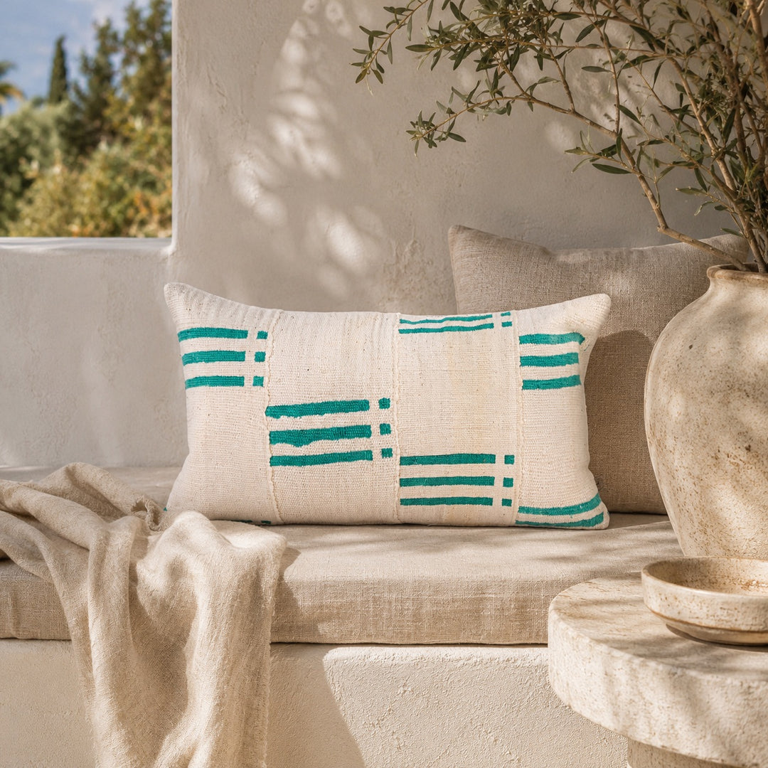 Housse de coussin NZETE par Studio Matongé en bogolan blanc et vert, mise en scène sur une banquette en lin dans un décor méditerranéen aux tons naturels.
