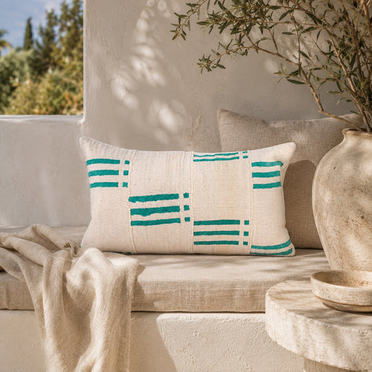 Housse de coussin NZETE par Studio Matongé en bogolan blanc et vert, mise en scène sur une banquette en lin dans un décor méditerranéen aux tons naturels.