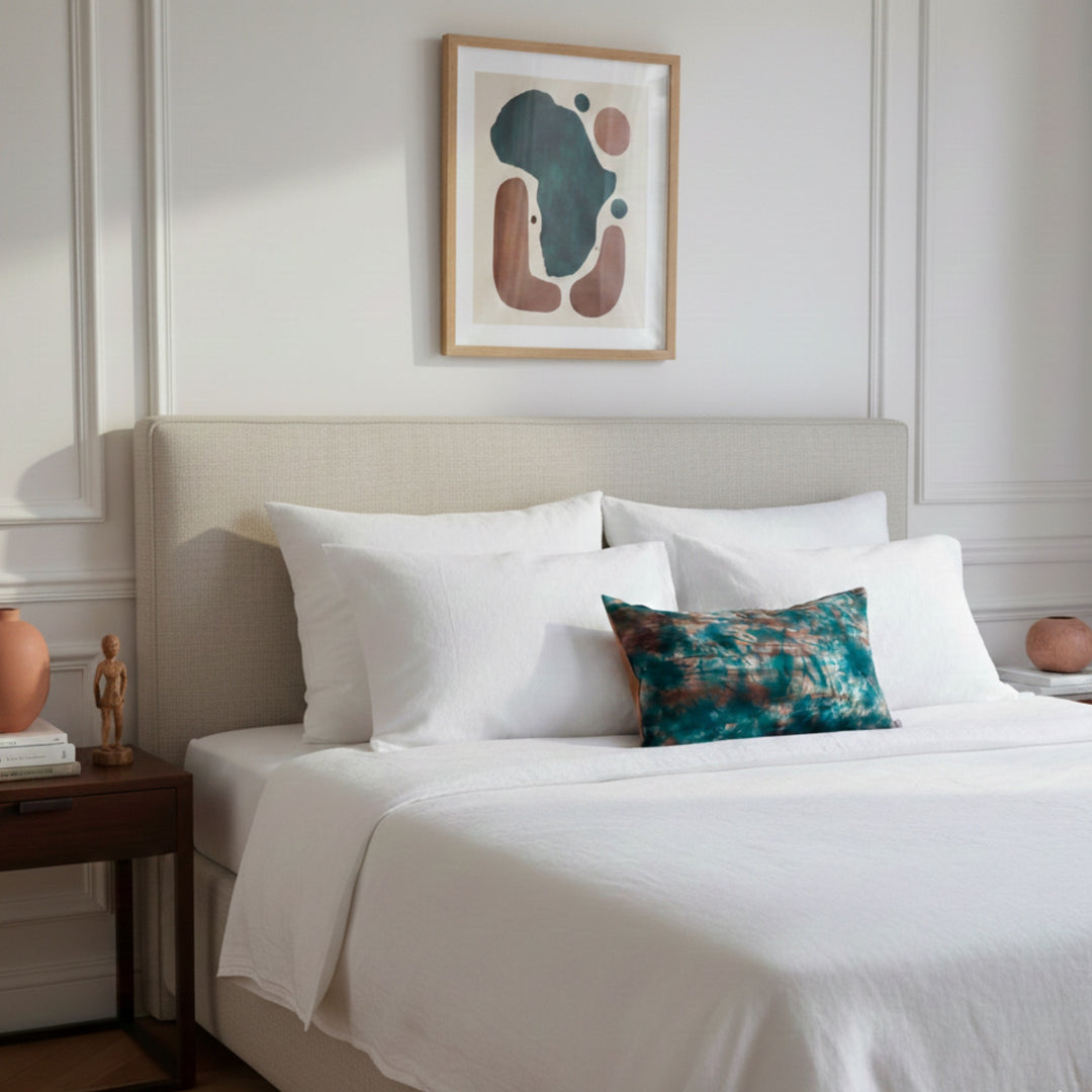 Housse de coussin KUMASI-LAGUNE posée sur un lit double aux draps blancs en lin, intérieur haussmannien lumineux, touche décorative textile artisanale.