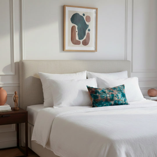 Housse de coussin KUMASI-LAGUNE posée sur un lit double aux draps blancs en lin, intérieur haussmannien lumineux, touche décorative textile artisanale.