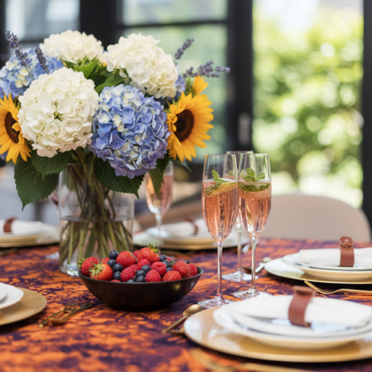 able dressée avec la nappe DUNDA – AMBER NIGHTS en kokodunda, vaisselle blanche, flûtes de champagne et centre de table floral, ambiance de repas lumineuse et estivale.