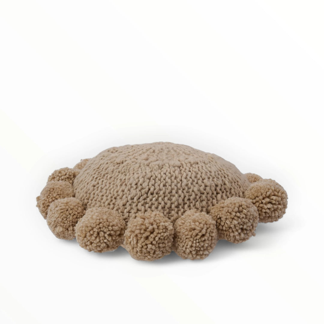 Coussin rond à pompons beige BONBONSI en pure laine mérinos, 30 cm de diamètre. Kenana Knitters pour Studio Matonge. Disponible en 2 tailles (30 cm et 50cm de diamètre) et 3 couleurs (blanc écru, beige et marron foncé). Insert fourni