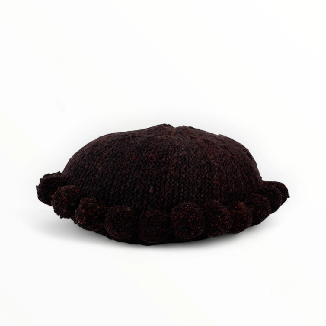 Coussin rond à pompons chocolat BONBONSI en pure laine mérinos, 50 cm de diamètre. Kenana Knitters pour Studio Matonge. Disponible en 2 tailles (30 cm et 50cm de diamètre) et 3 couleurs (blanc écru, beige et marron foncé). Insert fourni