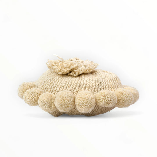 Coussin rond à pompons écru BONBONSI en pure laine mérinos, 30 cm de diamètre. Tricoté main par Kenana Knitters pour Studio Matongé. Disponible en 2 tailles (30 cm et 50cm de diamètre) et 3 couleurs (blanc écru, beige et marron foncé). Insert fourni