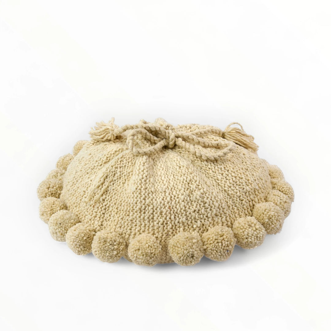 Coussin rond à pompons écru BONBONSI en pure laine mérinos, 50 cm de diamètre. Tricoté main par Kenana Knitters pour Studio Matongé. Disponible en 2 tailles (30 cm et 50cm de diamètre) et 3 couleurs (blanc écru, beige et marron foncé). Insert fourni