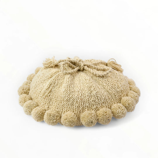 Coussin rond à pompons écru BONBONSI en pure laine mérinos, 50 cm de diamètre. Tricoté main par Kenana Knitters pour Studio Matongé. Disponible en 2 tailles (30 cm et 50cm de diamètre) et 3 couleurs (blanc écru, beige et marron foncé). Insert fourni