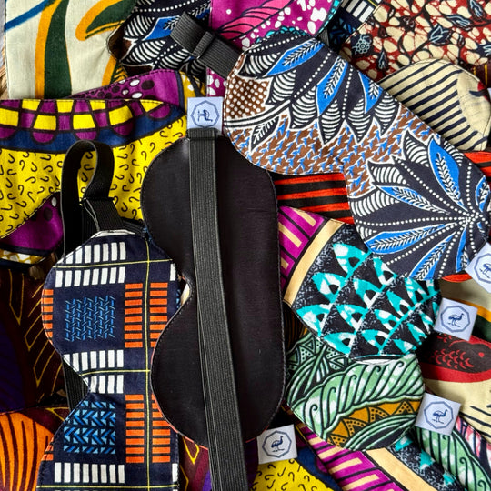 Masques de nuit DARE en wax et soie noire faits main par Studio Matongé, exposés en assortiment coloré – intérieur en soie côté paupières et élastique réglable visibles, confection artisanale en Belgique avec chutes de tissu upcyclées.