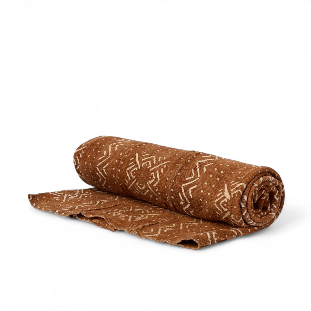 Plaid en bogolan de coton terracotta FEMI-ARDHI. Motifs ethniques écrus. En coton bio filé, tissé, teint et décoré à la main par nos artisans dans la région de Ségou au Mali. Approximativement 153 x 117 cm.