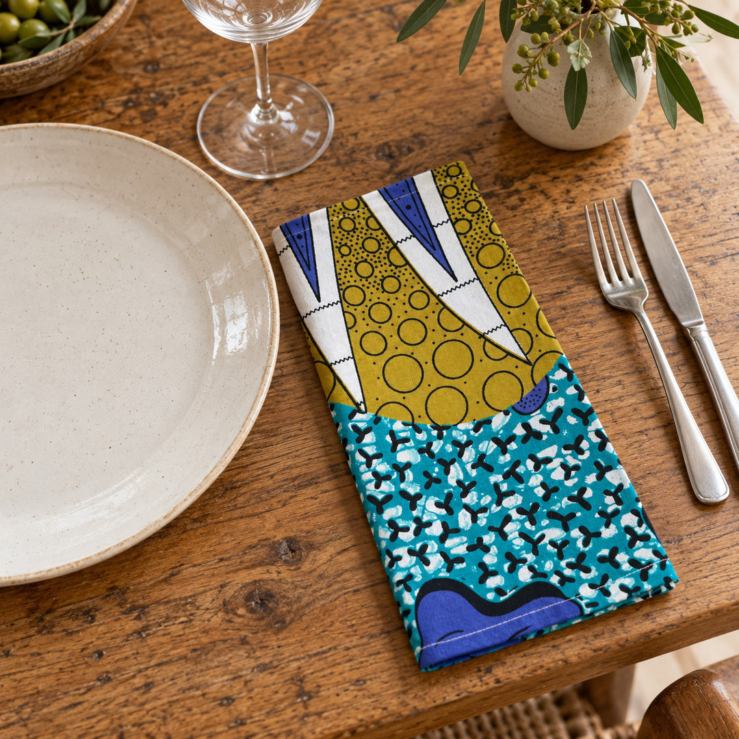 Serviette de table AYO-LAGA pliée à côté d’une assiette en céramique écrue sur une table en bois.