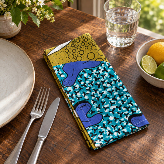 Serviette de table AYO-LAGA sur une table de déjeuner en bois, près d’une assiette claire, d’un verre et d’un bol d’agrumes.