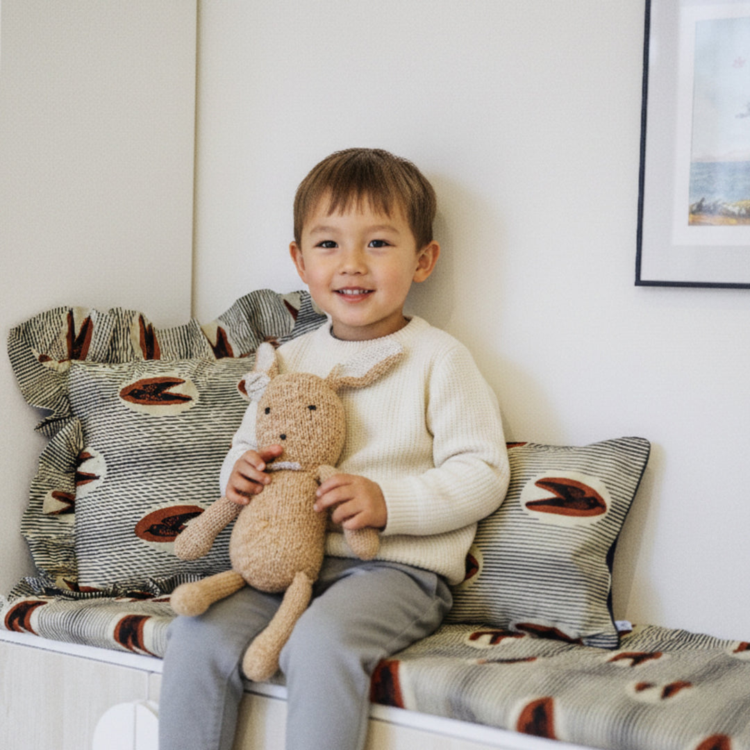 Enfant assis sur une banquette, tenant un doudou en laine SIMONE, entouré de housses de coussin en wax LONA-MTOTO et FEFERE-MTOTO à volants, vue d’ensemble.