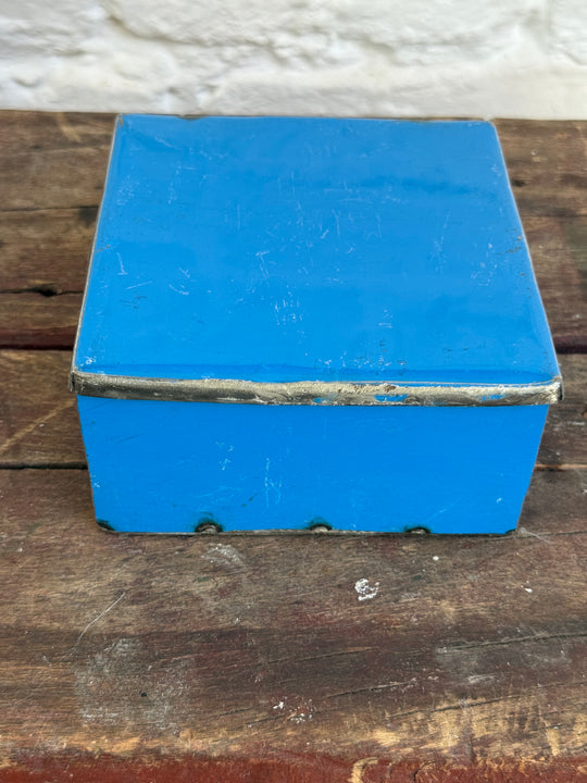 Boîte RANGI bleue en métal recyclé, couvercle fermé, vue de face sur un tabouret en bois.