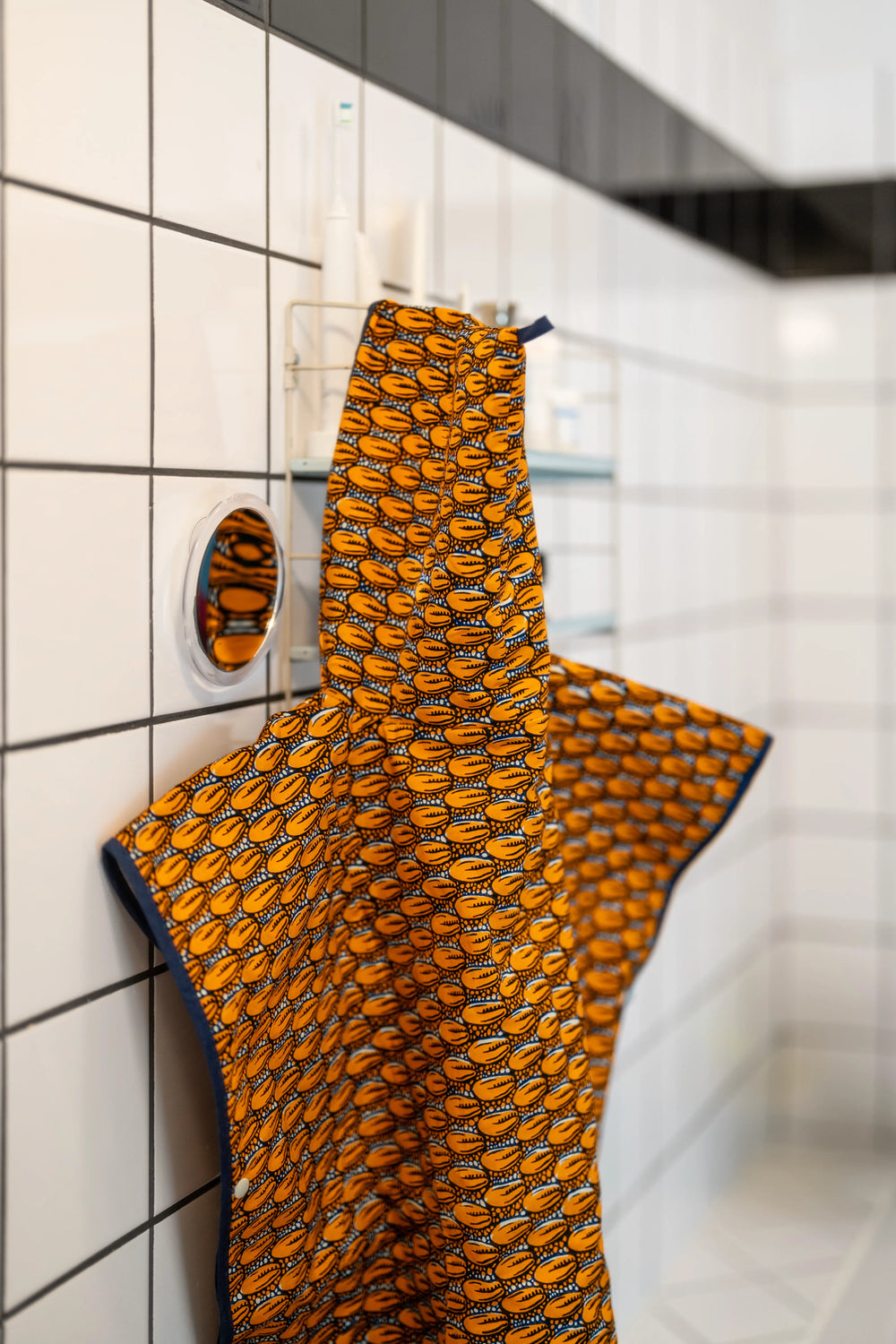 Poncho de bain enfant SIMBA-CAURI en wax orange à cauris suspendu près d'un miroir dans une salle de bain carrelée.