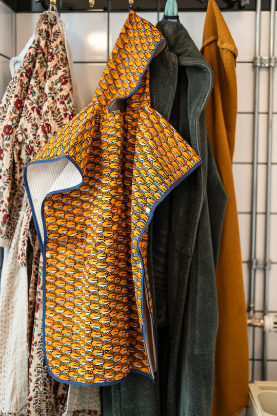 Poncho de bain enfant SIMBA-CAURI en wax orange à motif cauris, biais bleu marine et envers éponge, suspendu à une patère