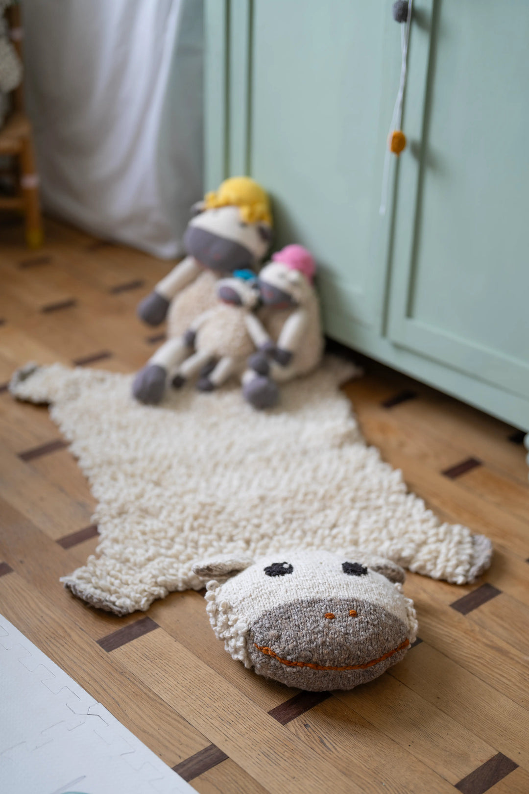 Dans une chambre d'enfant aux tons pastels, le tapis mouton SHEEPY, tricoté main en pure laine, et la famille de doudous moutons HECTOR en coton bio (disponibles en 3 tailles, couleur du chapeau aléatoire)