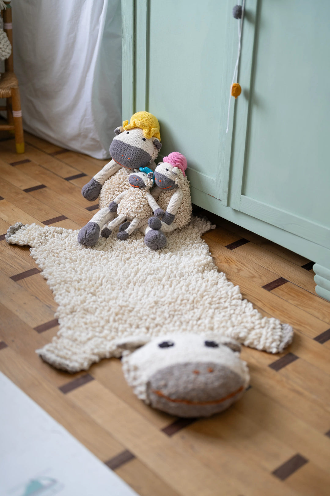 Dans une chambre d'enfant aux tons pastels, le tapis mouton SHEEPY, tricoté main en pure laine, et la famille de doudous moutons HECTOR en coton bio (disponibles en 3 tailles, couleur du chapeau aléatoire)