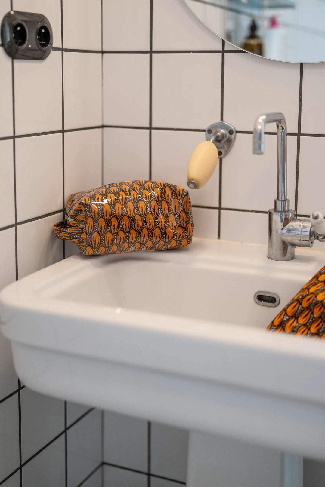 Trousse étanche ZAZA-CAURI en wax orange à cauris posée près d'un robinet chromé, un angle plongeant dans le lavabo.