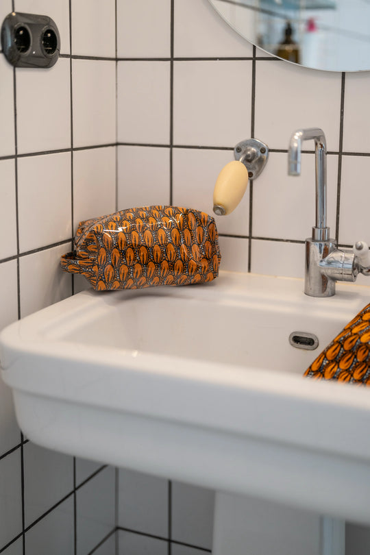 Trousse étanche ZAZA-CAURI en wax orange à cauris posée près d'un robinet chromé, un angle plongeant dans le lavabo.