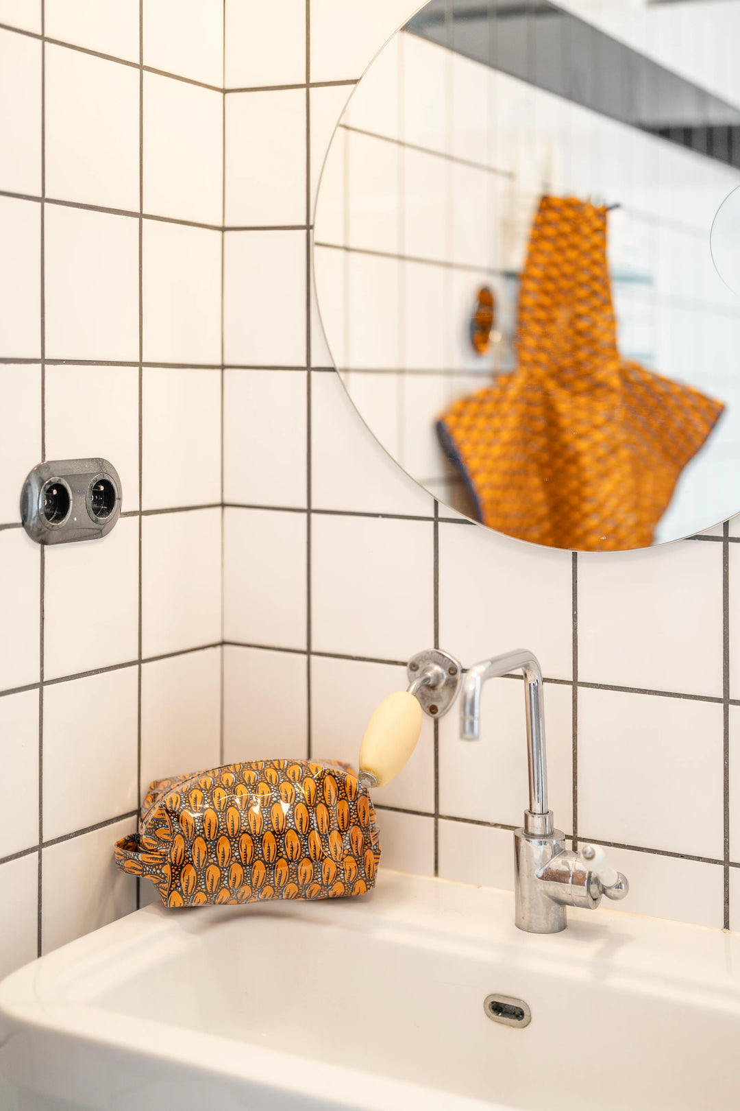 Trousse de toilette étanche ZAZA-CAURI en wax orange à cauris posée sur le rebord d'un lavabo en faïence blanche.