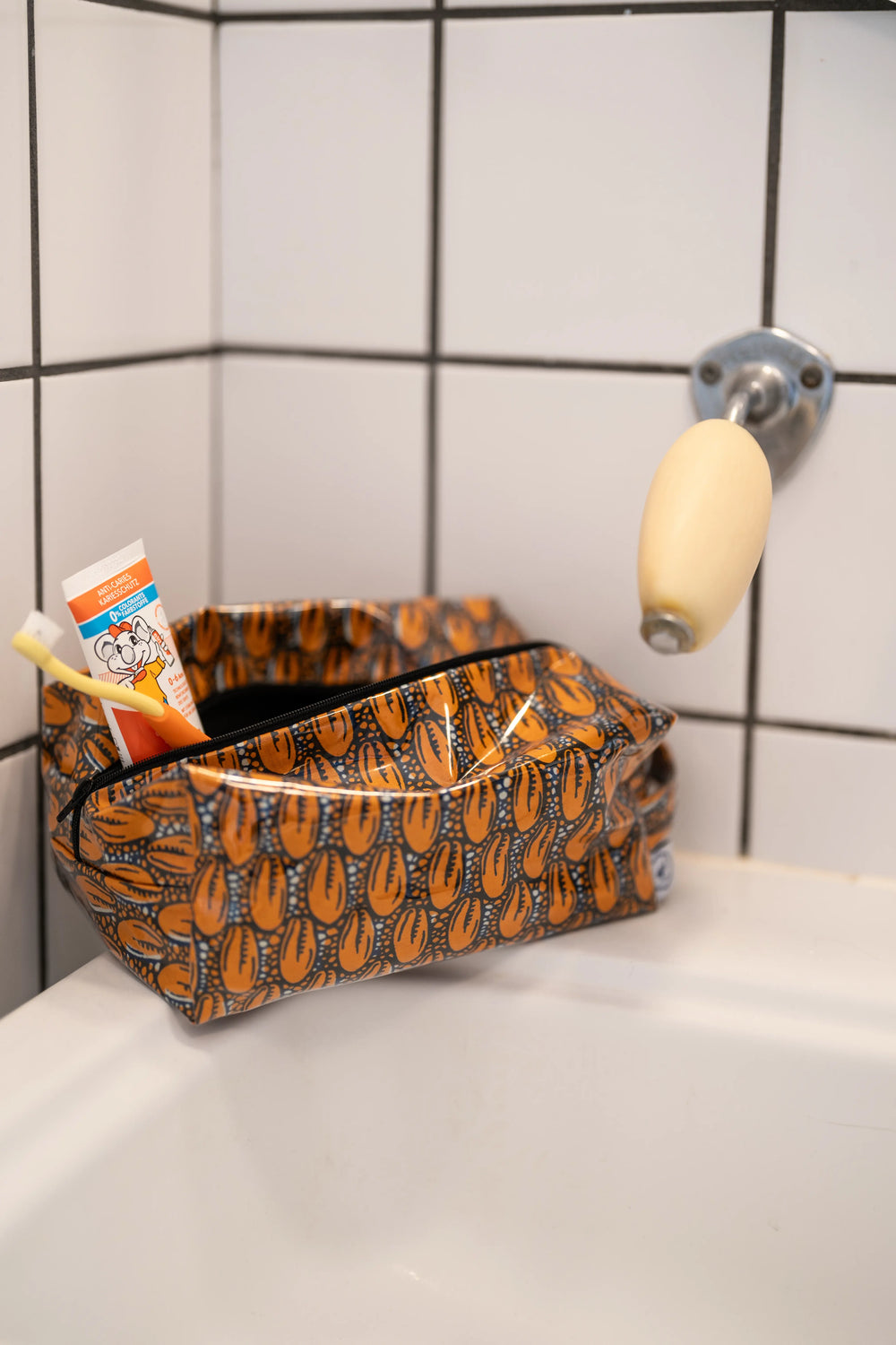Trousse étanche ZAZA-CAURI en wax orange à cauris posée près d'un lavabo carrelé, reflétée dans le miroir rond.