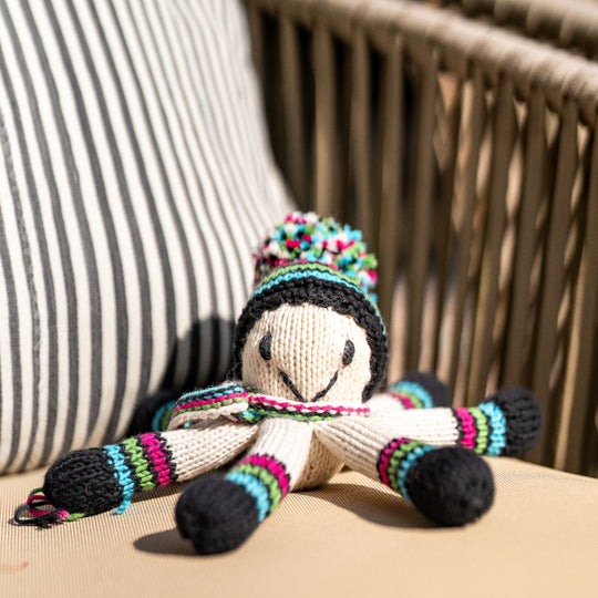 Doudou pieuvre NAIA en laine tricotée main, bonnet à pompon multicolore et tentacules rayées turquoise, vert noir et fuschia, posé sur un fauteuil en rotin.