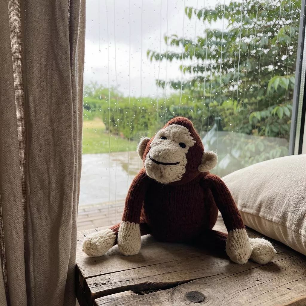 André, doudou singe en laine mérinos tricoté à la main, est assis sur un banc en bois brut devant la fenêtre.