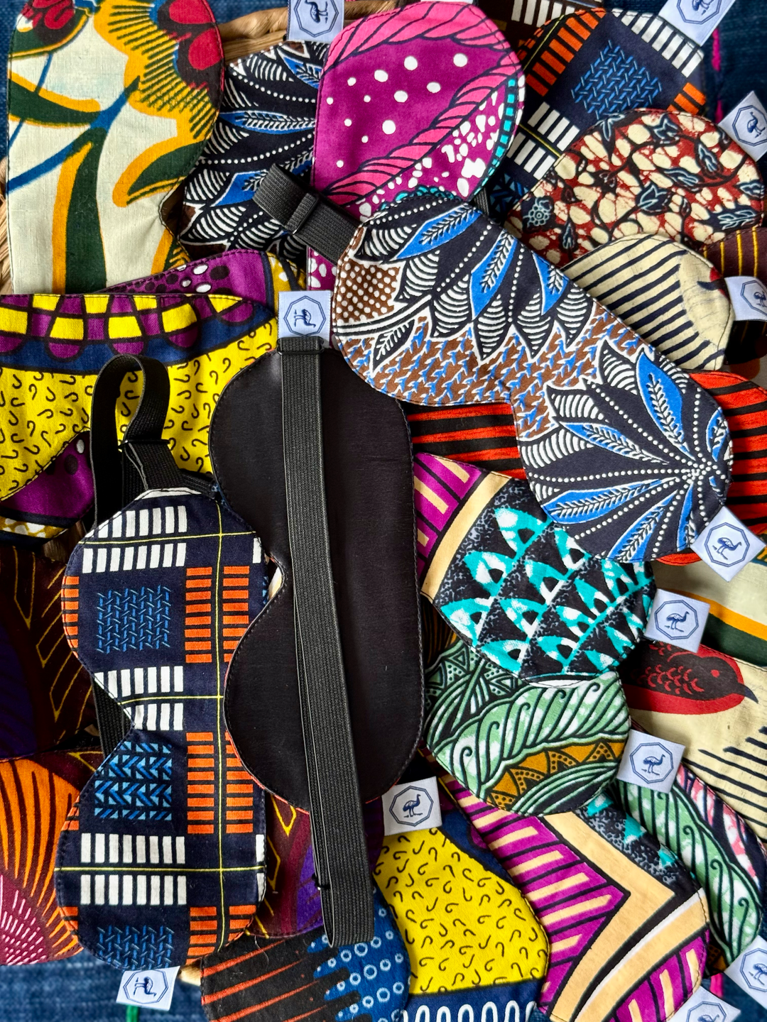 Masques de nuit DARE en wax et soie noire faits main par Studio Matongé, exposés en assortiment coloré – intérieur en soie côté paupières et élastique réglable visibles, confection artisanale en Belgique avec chutes de tissu upcyclées.