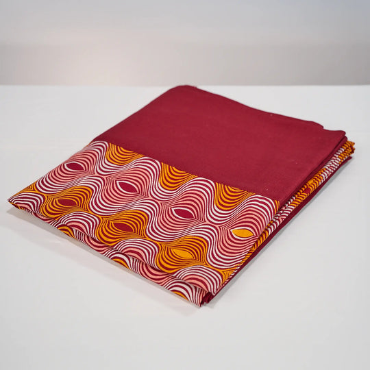 Nappe GIDA_ZUBA pliée sur fond blanc. Bord en lin bordeaux et centre en wax bordeaux, abricot, rose dragée et blanc.