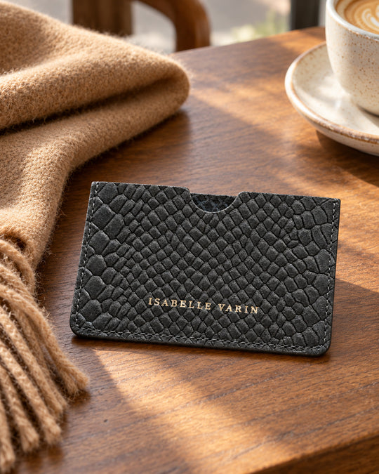 CREDY-Kartenetui von Isabelle Varin aus schokoladenbraunem Nubukleder mit Kroko-Prägung, auf einem Holztisch neben einem Schal und Kaffee.